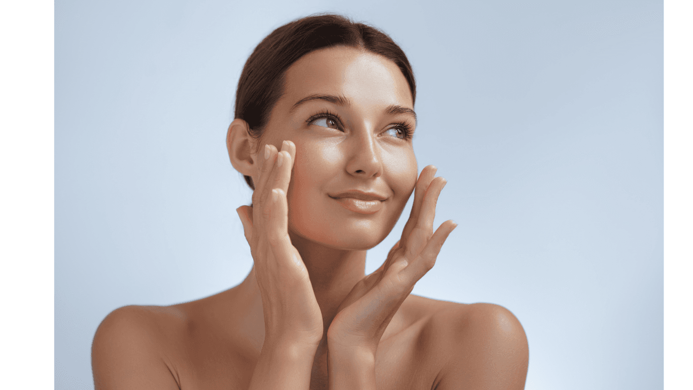 A woman with radiant skin framing her face with her hands in soft light.
