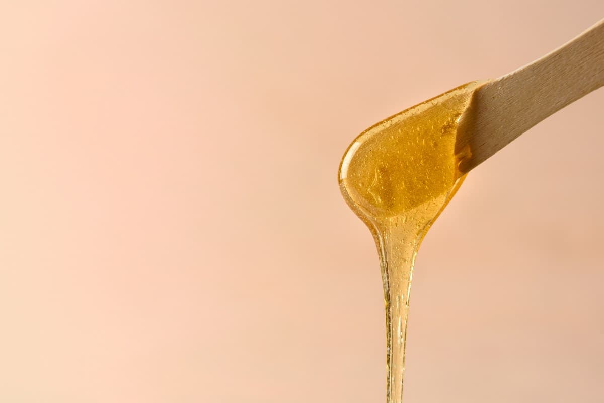 Warm golden wax drizzling from a wooden spatula against a soft peach backdrop.